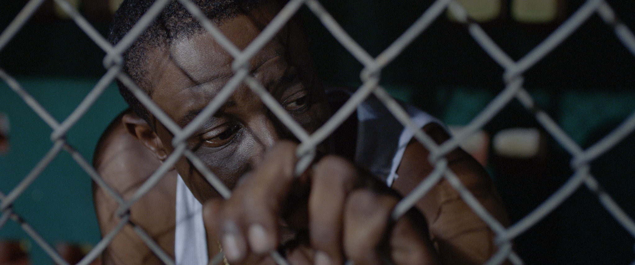 A man’s face pressed close behind chain-link fencing, eyes lowered.