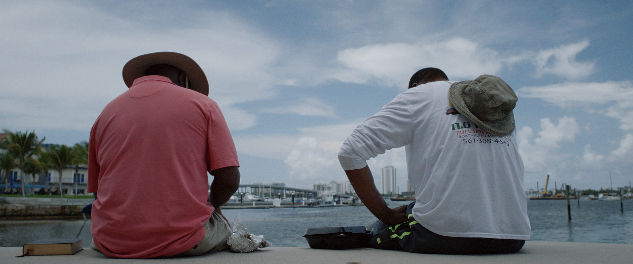 Two men sit at the edge of a dock looking out over open water on an overcast afternoon.