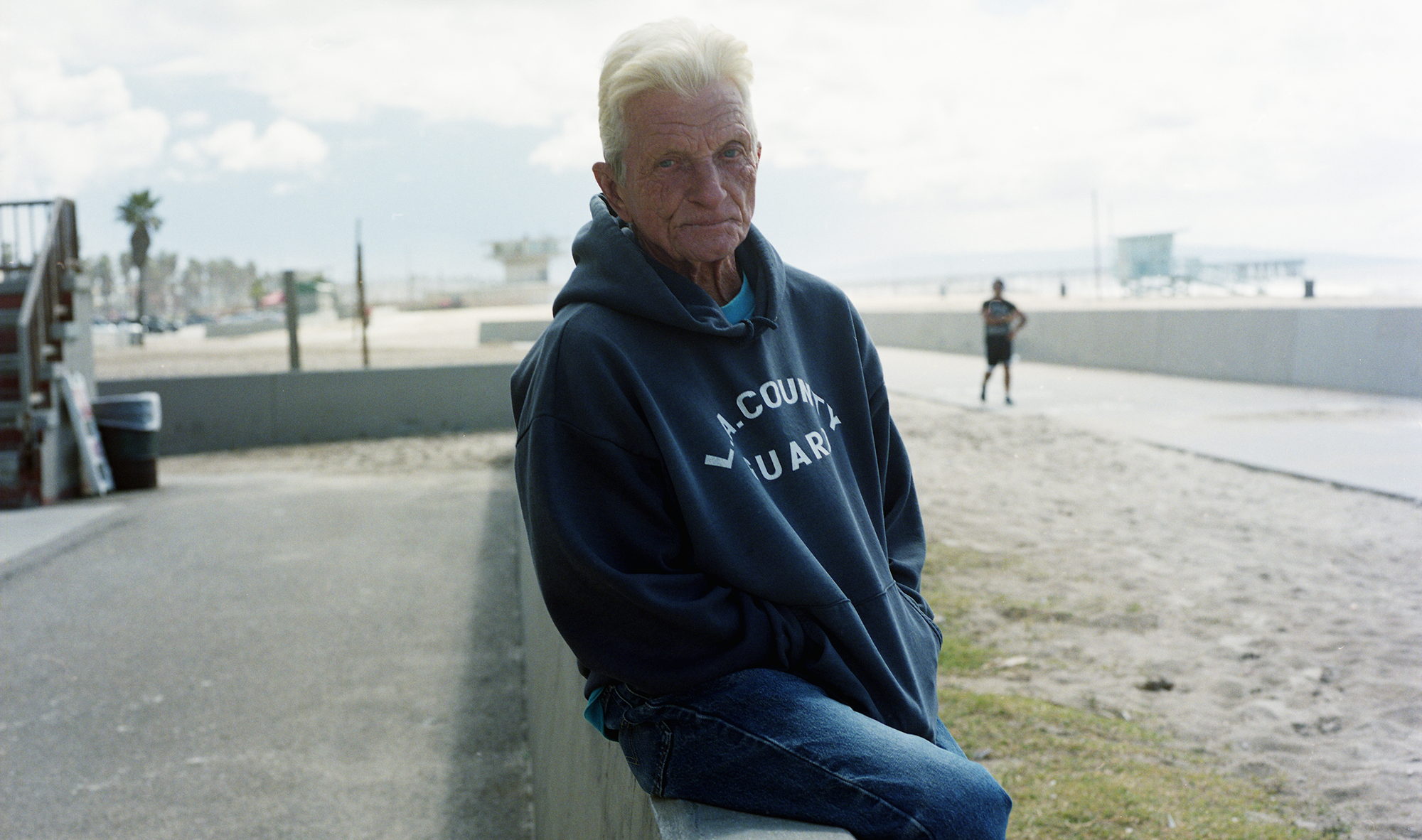 An older man in a hooded sweatshirt rests against a low seawall on a wide stretch of sand; a lone runner passes far behind him, palms and a lifeguard tower on the horizon.