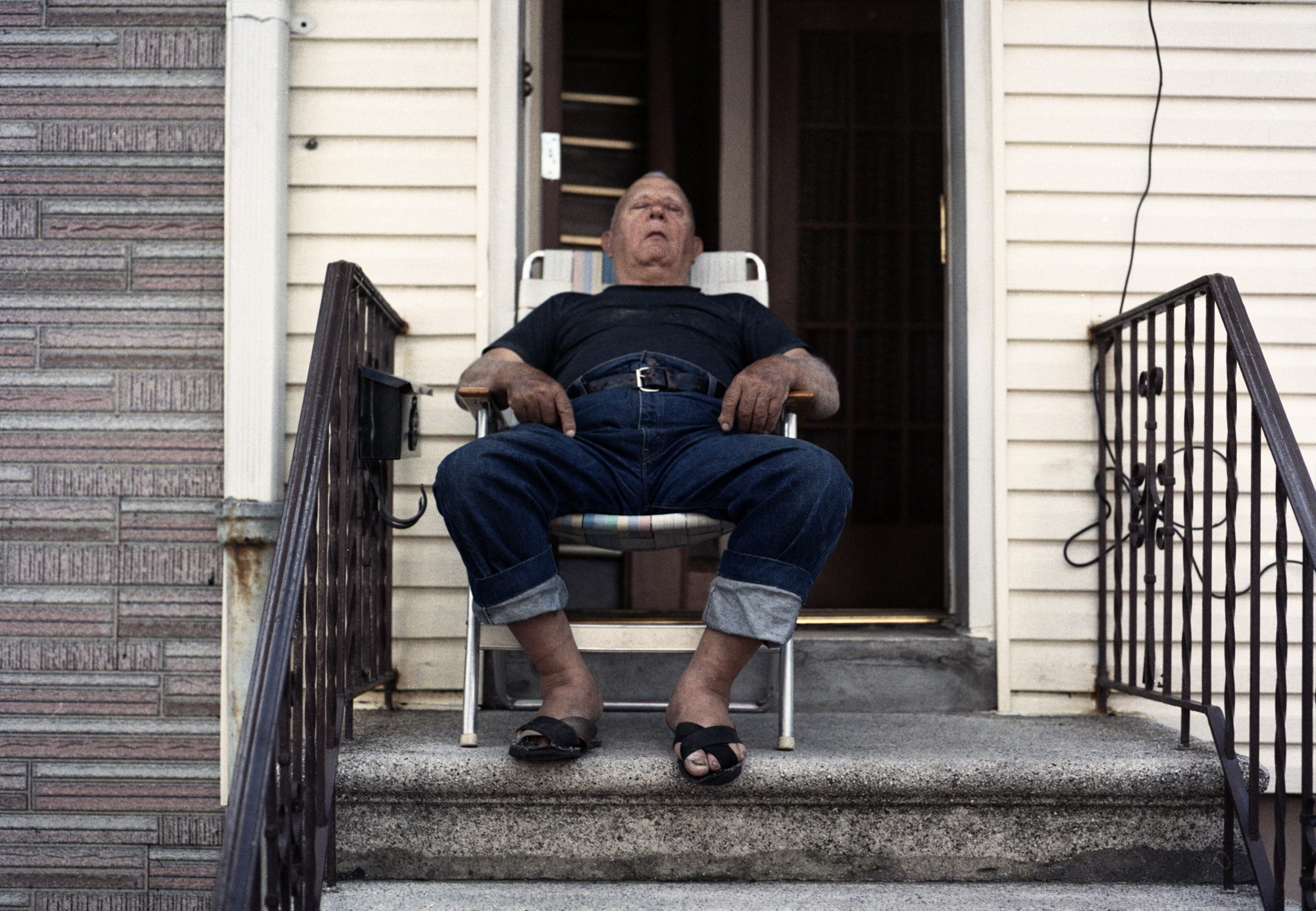 A heavy-set man sits back in a folding chair in the open doorway of a clapboard house, eyes closed against the sun.