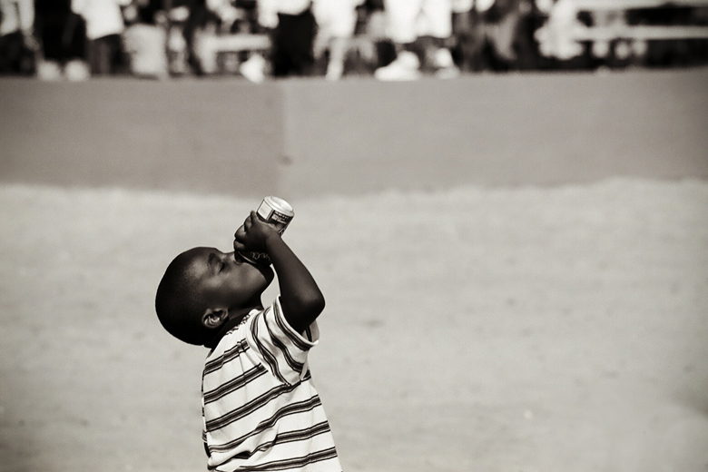 A small boy tips a bottle back to drink, a blurred crowd behind him.