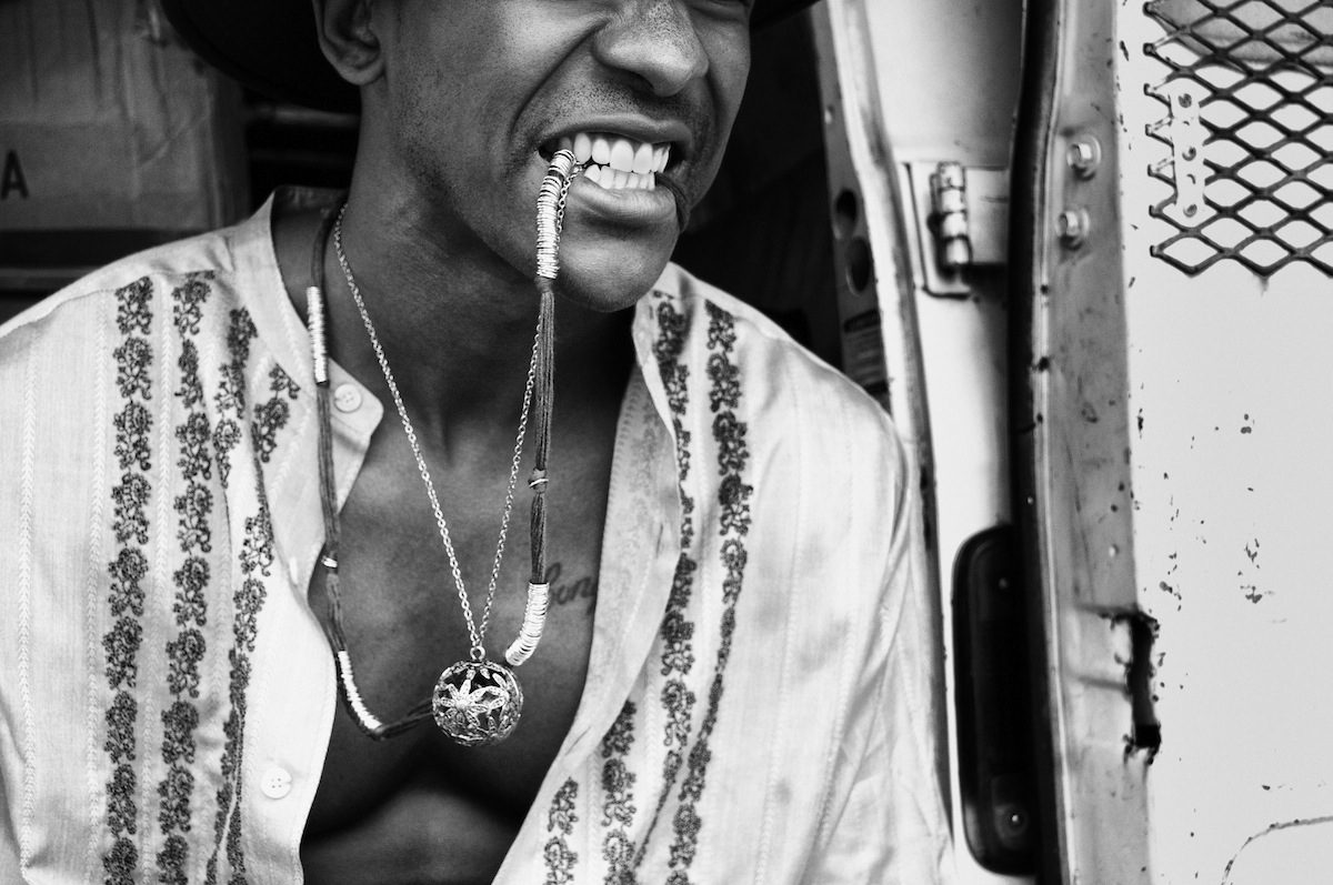 A young man bites down on a chain, an open embroidered shirt at his chest, the side of a van behind him.