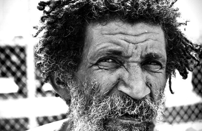 Close portrait of an older man with long grey locs and a full beard, looking directly into the camera.