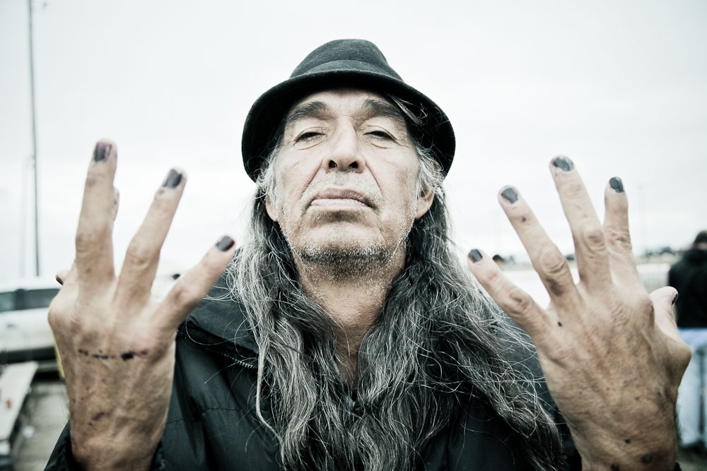 A long-haired man in a hat raises both hands toward the camera, fingernails painted dark, boats behind him.