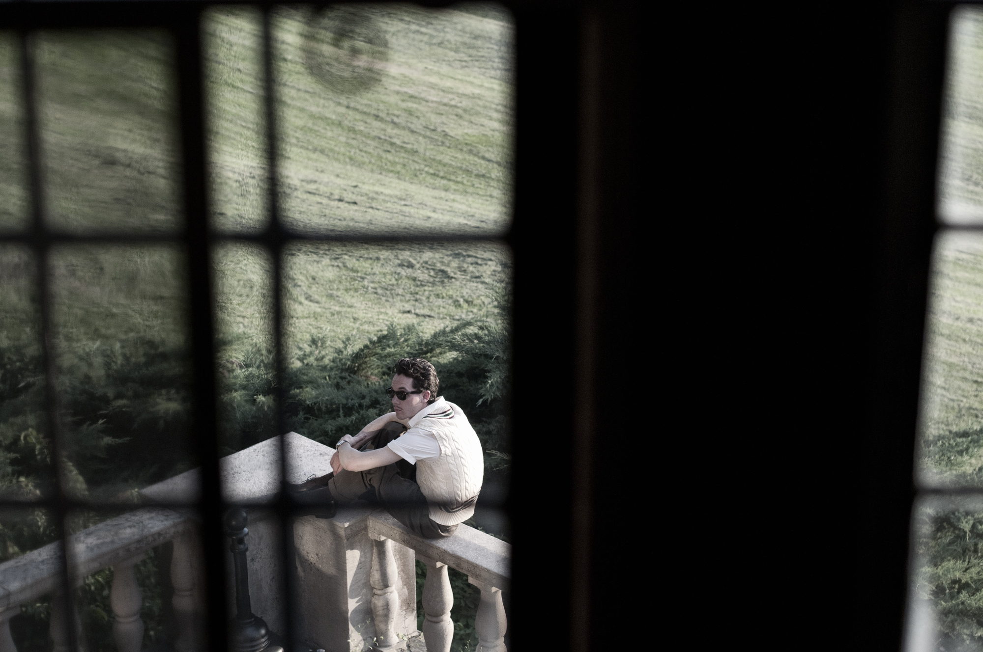 Seen through the panes of a tall window, a man sits alone on a balustrade above a lawn.