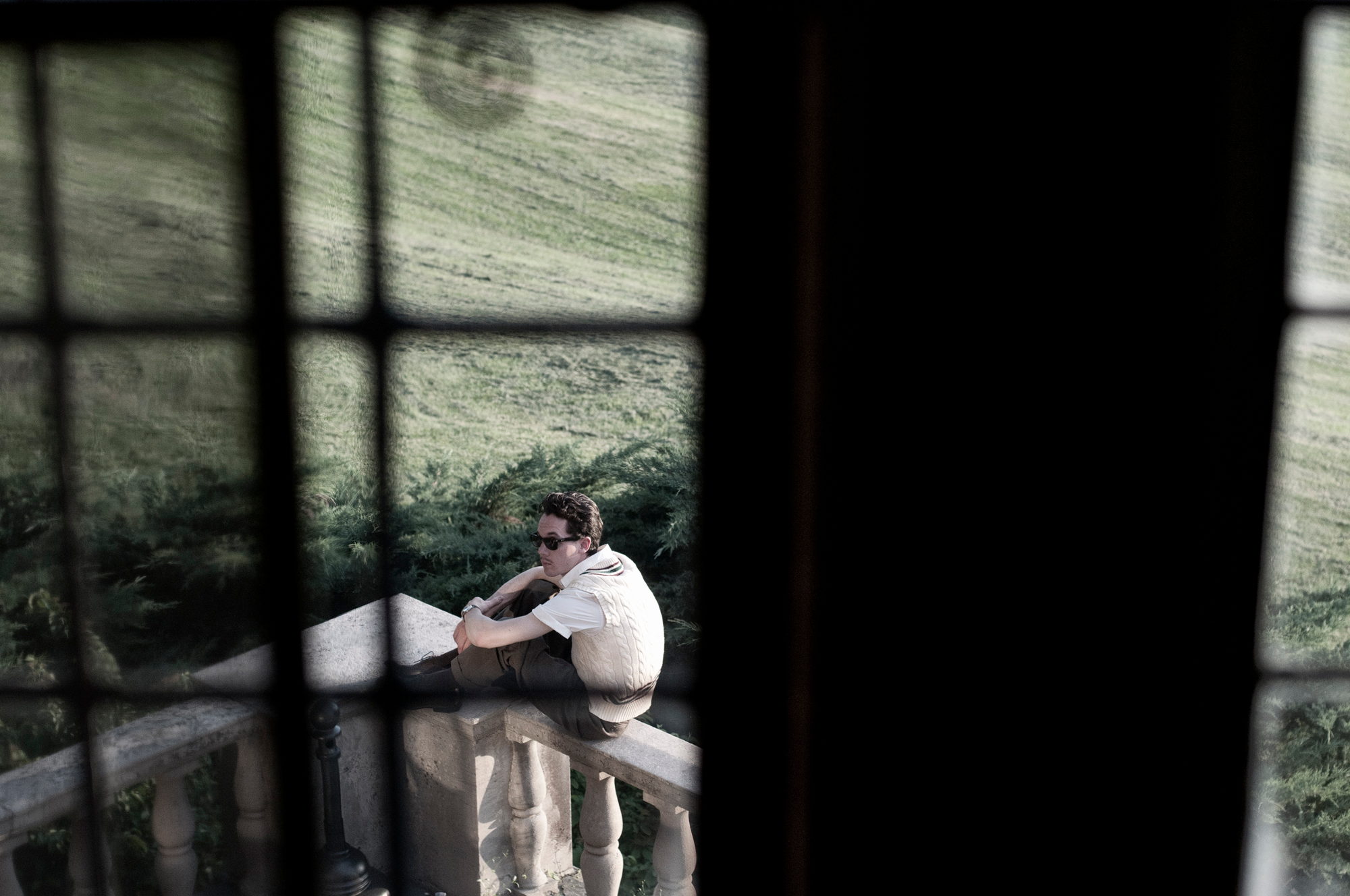 Seen through the panes of a tall window, a man sits alone on a balustrade above a lawn.