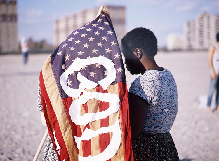 On a beach, a woman stands beside an American flag with the word GOD painted across it; apartment towers rise behind.