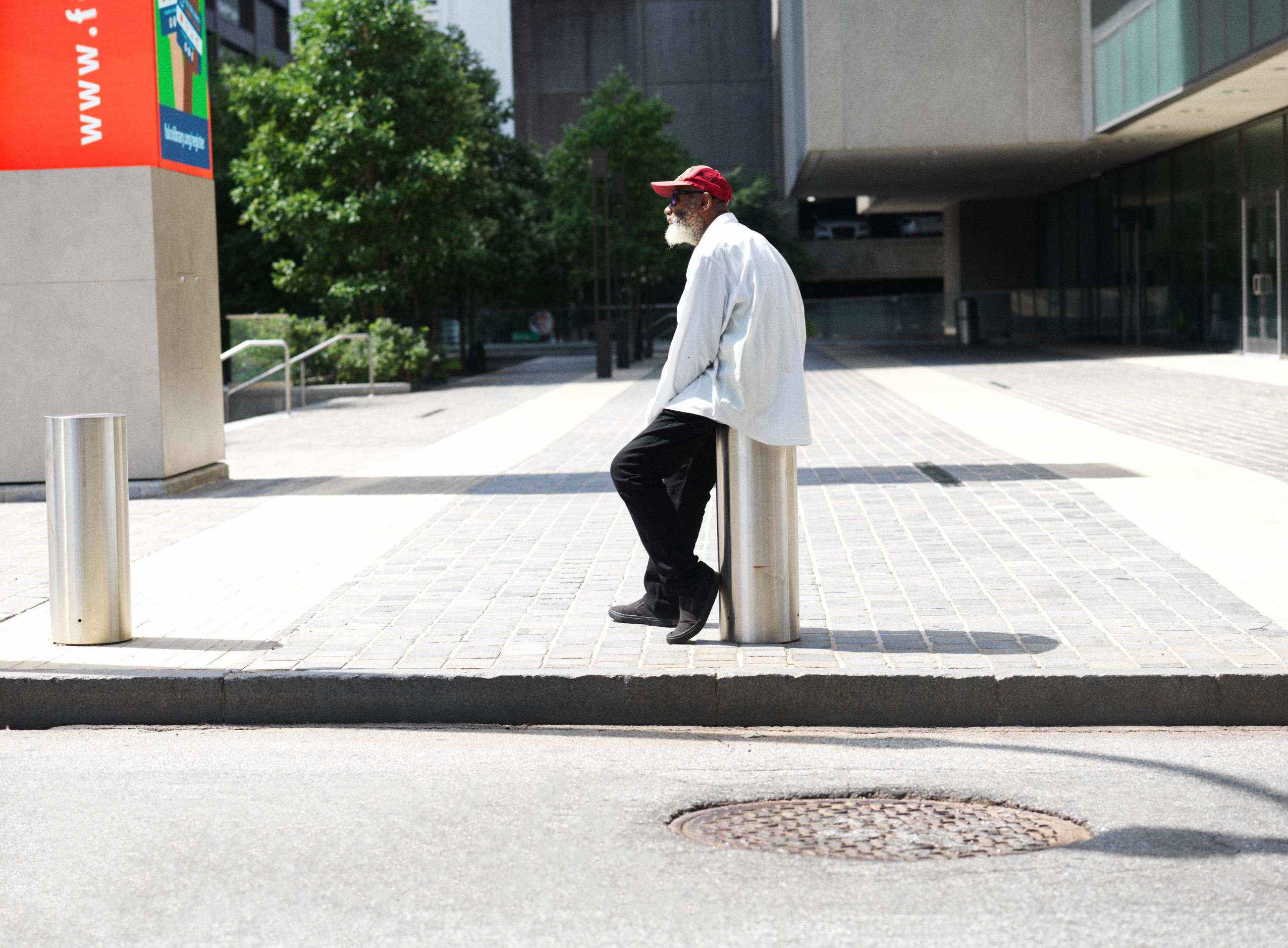 An older man in a red cap and a loose white shirt rests back against a steel bollard on an empty paved plaza, alone in hard midday light between office buildings.
